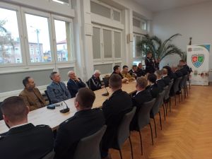 Na zdjęciu znajdują się strażacy siedzący przy stole, jeden z nich zabiera głos. Wśród strażaków znajduje się również...